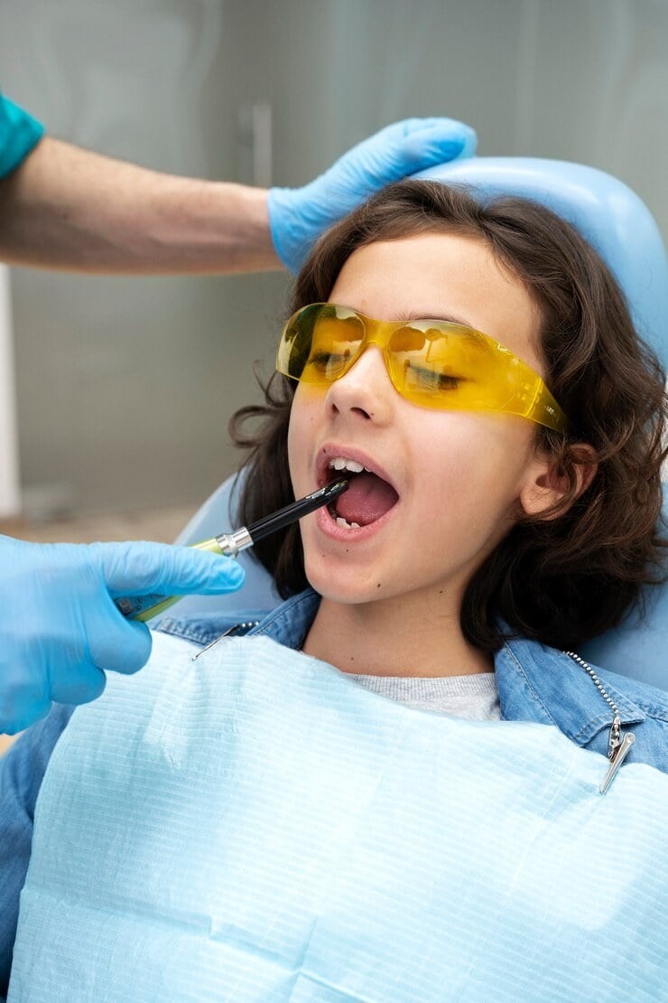 A child in yellow safety glasses having a dental check-up, with a gloved hand holding a dental instrument