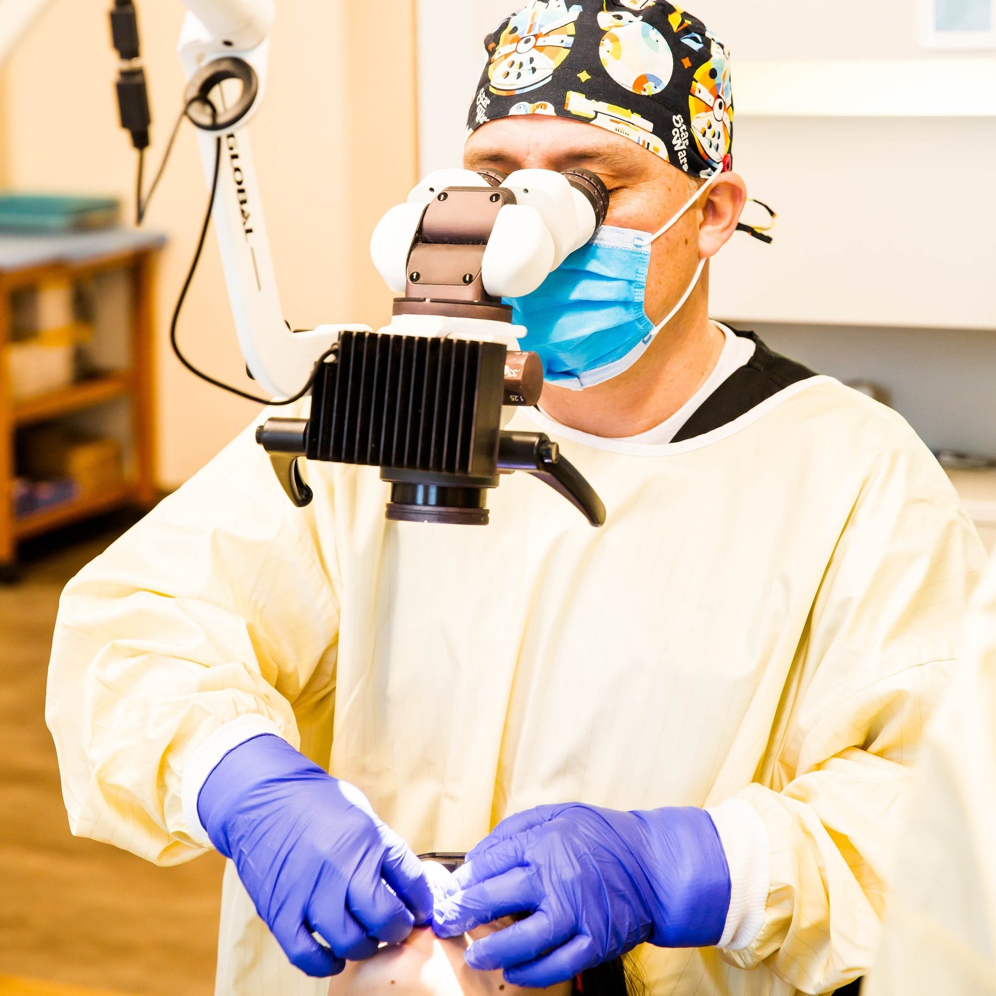 A dentist in a mask and protective gown working at a dental microscope