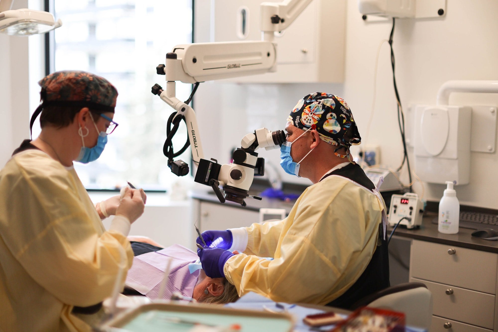 A clinician in a patterned cap, mask and yellow gown working at a dental microscope